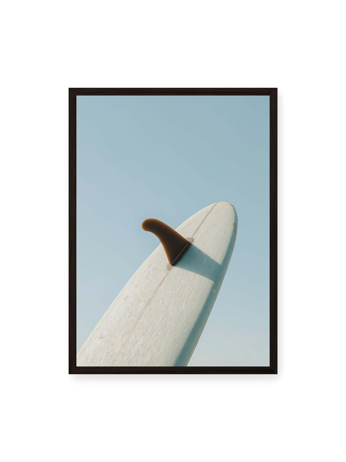 50x70 cm, schwarzer Rahmen – Helles Surfbrett mit dunkler Finne und markantem Schatten ragt diagonal vor einen klaren blauen Himmel.