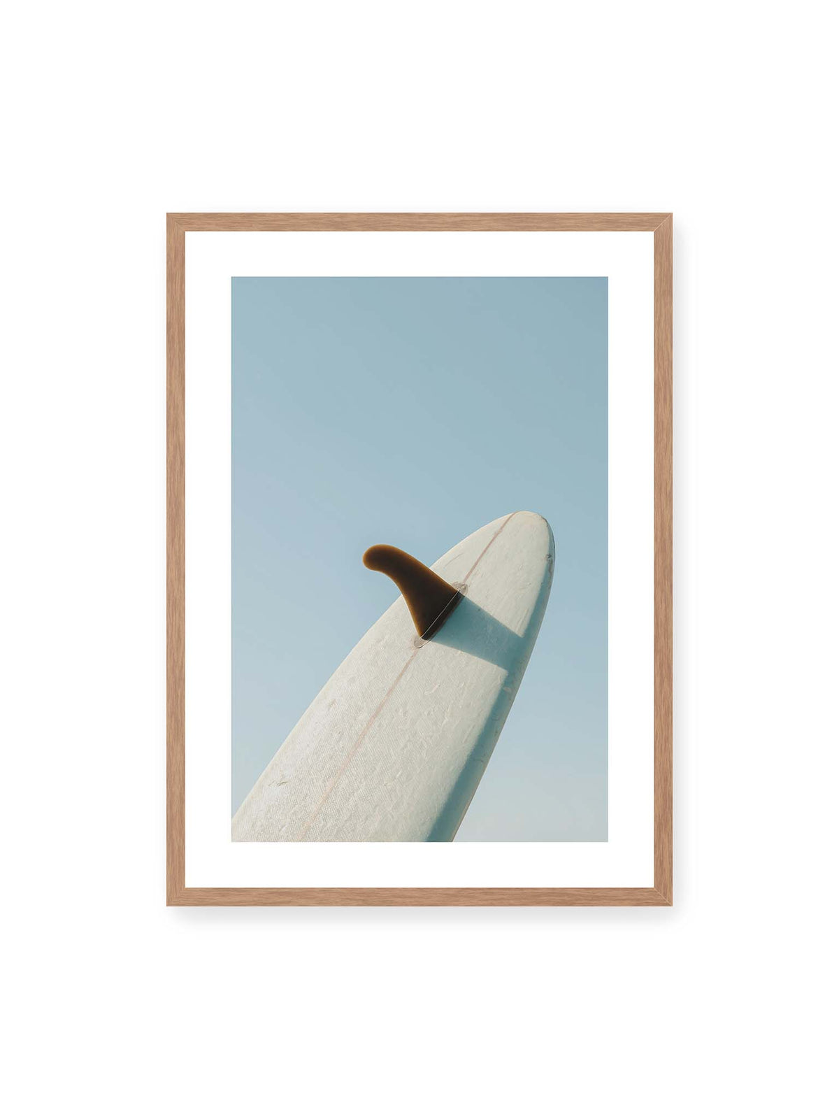 50x70 cm, weißer Galerie-Rand, Eichenrahmen – Helles Surfbrett mit dunkler Finne und markantem Schatten ragt diagonal vor einen klaren blauen Himmel.