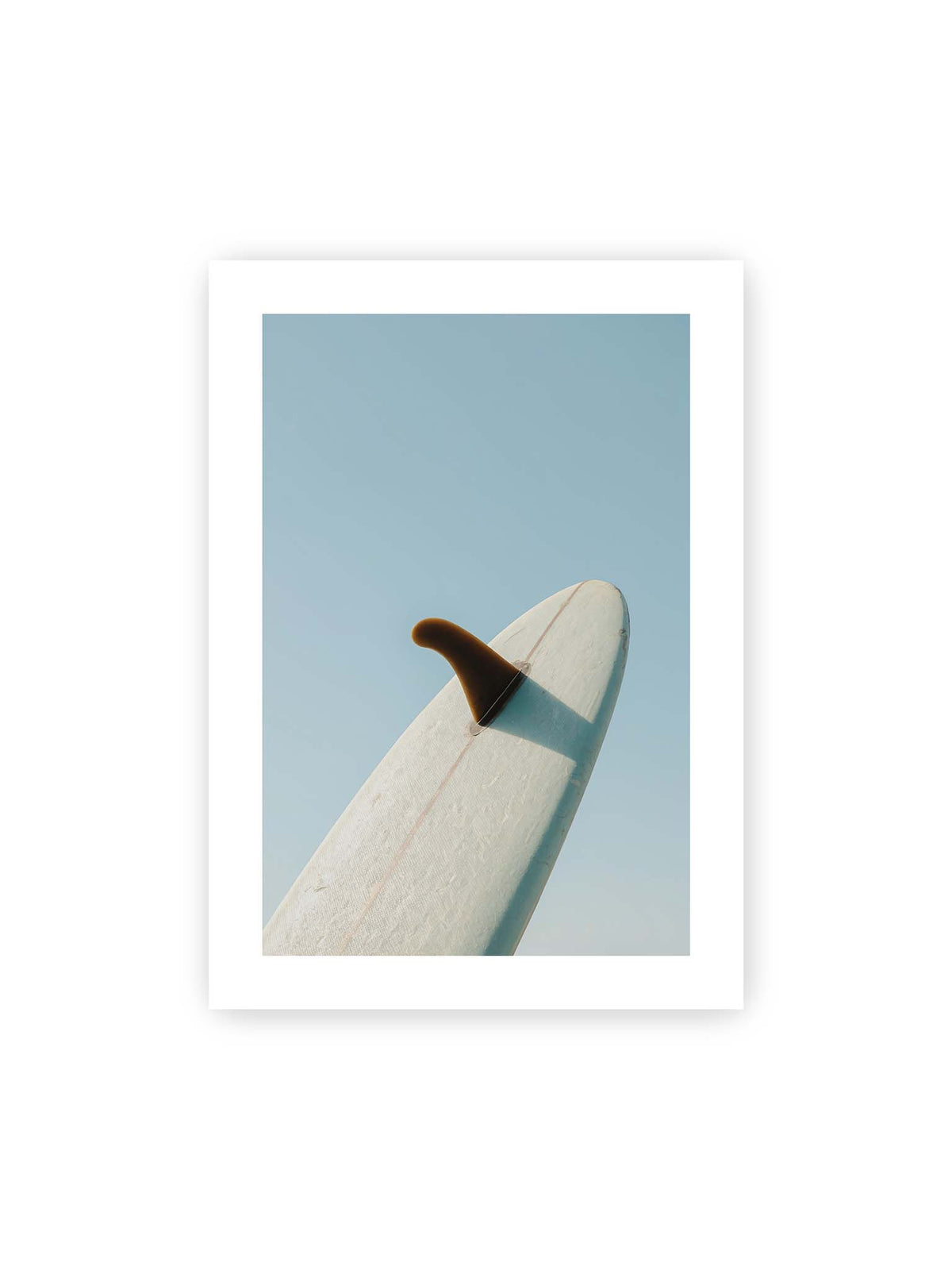 50x70 cm, weißer Galerie-Rand – Helles Surfbrett mit dunkler Finne und markantem Schatten ragt diagonal vor einen klaren blauen Himmel.