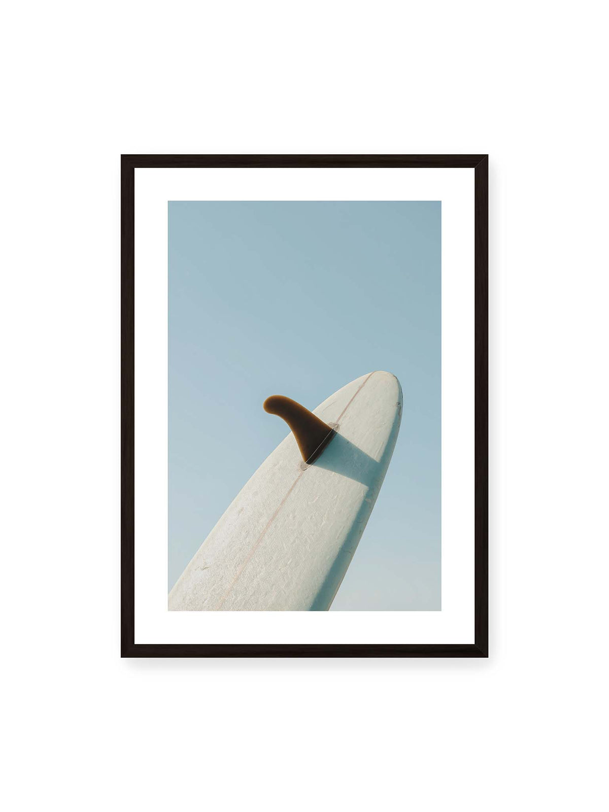 50x70 cm, weißer Galerie-Rand, schwarzer Rahmen – Helles Surfbrett mit dunkler Finne und markantem Schatten ragt diagonal vor einen klaren blauen Himmel.
