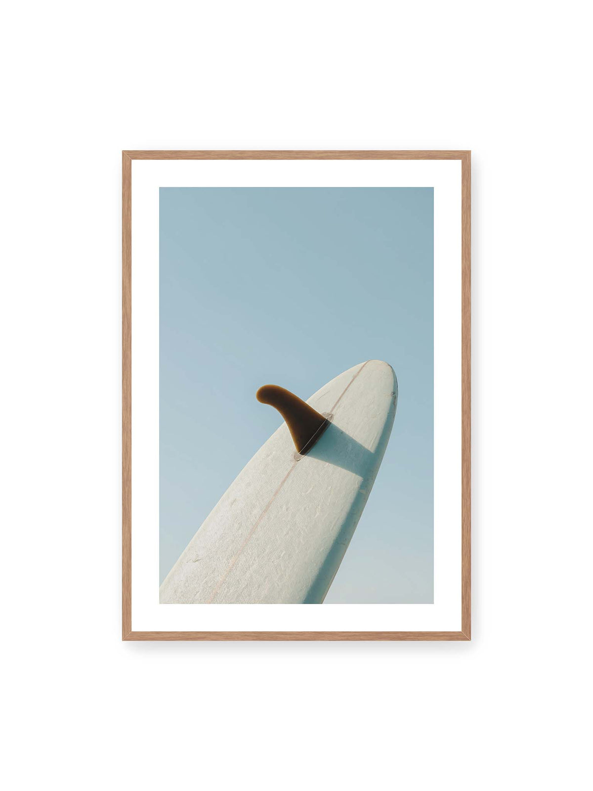 70x100 cm, weißer Galerie-Rand, Eichenrahmen – Helles Surfbrett mit dunkler Finne und markantem Schatten ragt diagonal vor einen klaren blauen Himmel.