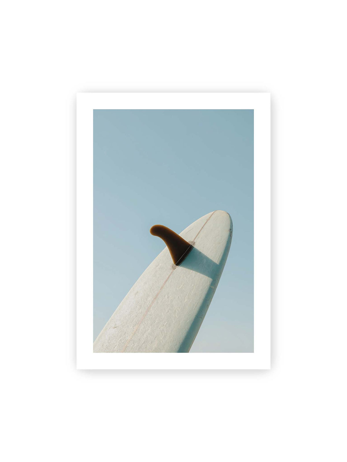 70x100 cm, weißer Galerie-Rand – Helles Surfbrett mit dunkler Finne und markantem Schatten ragt diagonal vor einen klaren blauen Himmel.