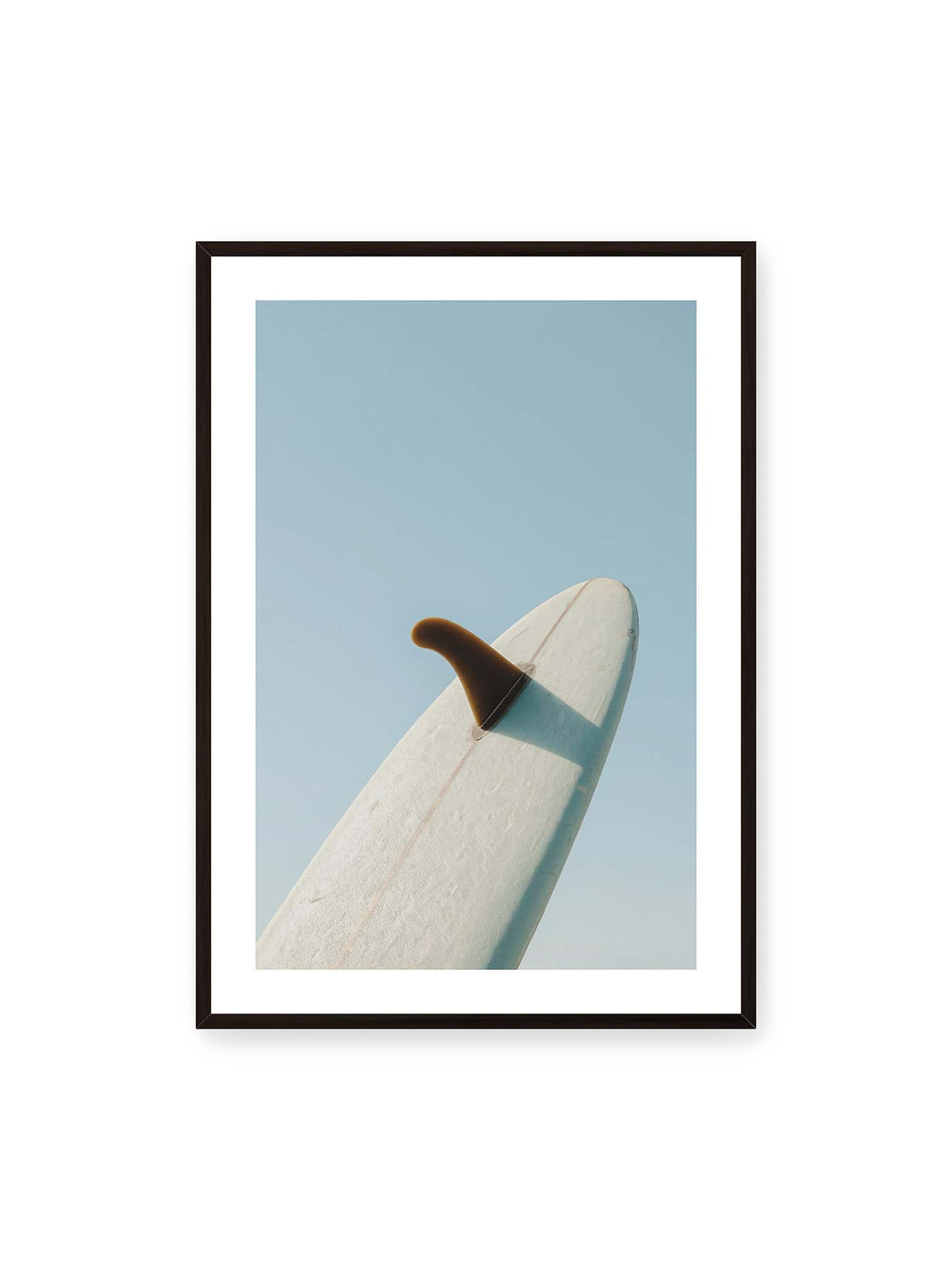 70x100 cm, weißer Galerie-Rand, schwarzer Rahmen – Helles Surfbrett mit dunkler Finne und markantem Schatten ragt diagonal vor einen klaren blauen Himmel.