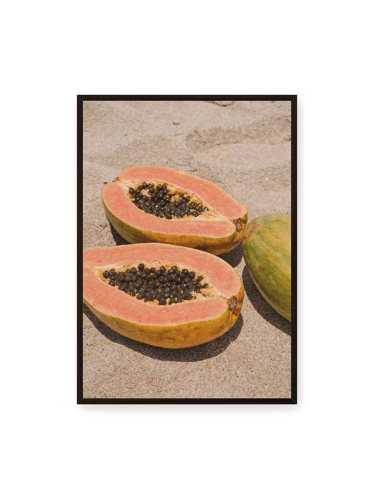 70x100 cm, schwarzer Rahmen – Zwei geöffnete Papayahälften mit dunklen Samen und eine ganze Papaya auf hellem Sand.